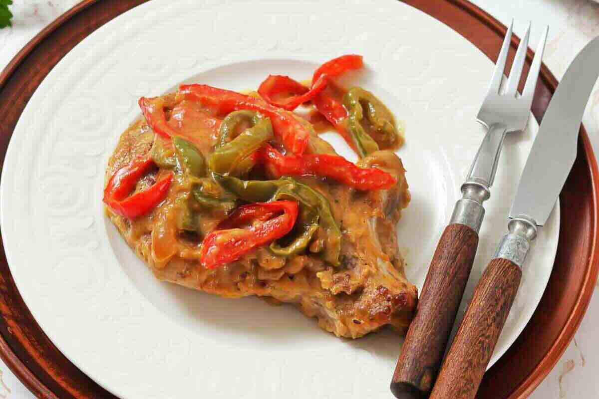 Pork chop topped with cooked red and green bell peppers and sauce, served on a white plate with a fork and knife.