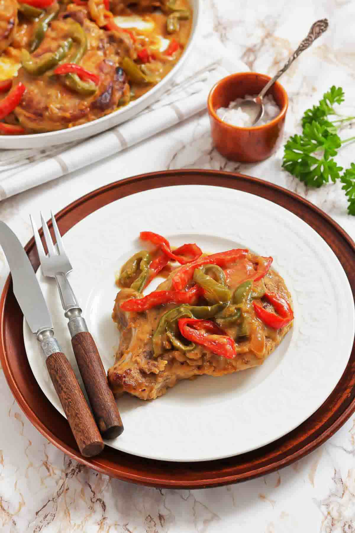Cooked pork chop topped with sautéed red and green bell peppers on a white plate, with a fork and knife beside it and a pan of food in the background.