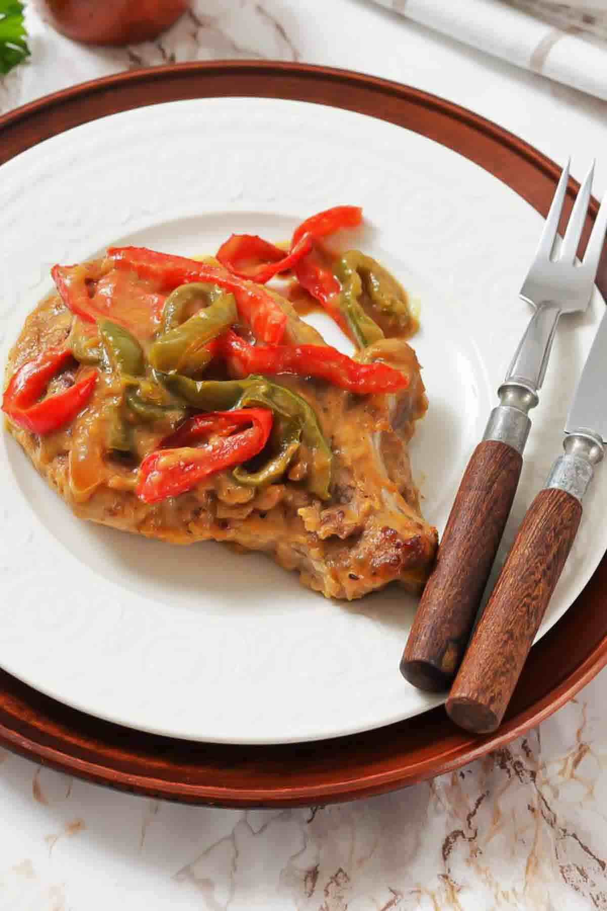 A cooked pork chop topped with sautéed red and green bell peppers and sauce, served on a white plate with a fork and knife on the side.
