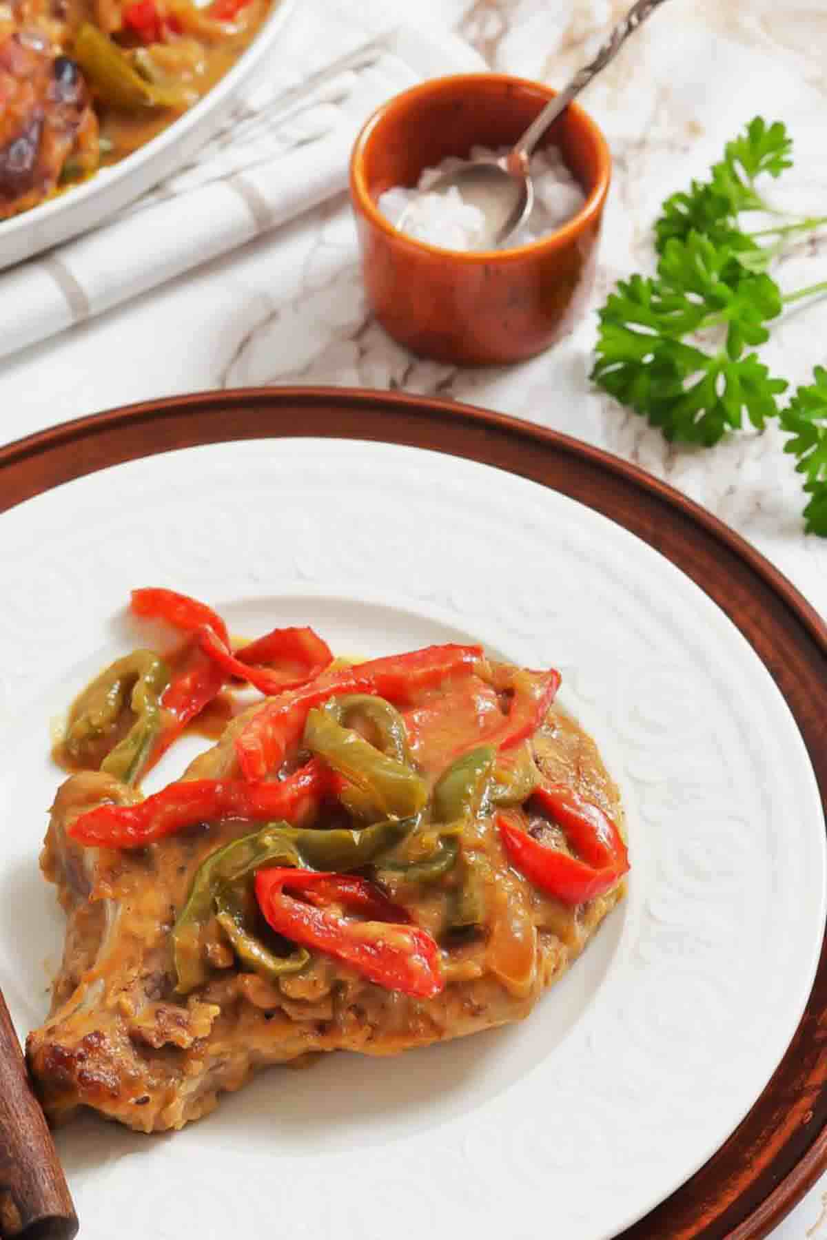 A cooked pork chop topped with sautéed red and green bell peppers and creamy sauce, served on a white plate with a brown rim. Parsley and a small bowl of salt are in the background.