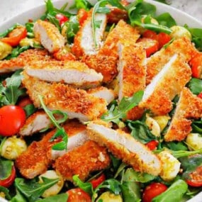 A plate of salad with arugula, cherry tomatoes, mozzarella balls, and sliced breaded chicken cutlets on top.