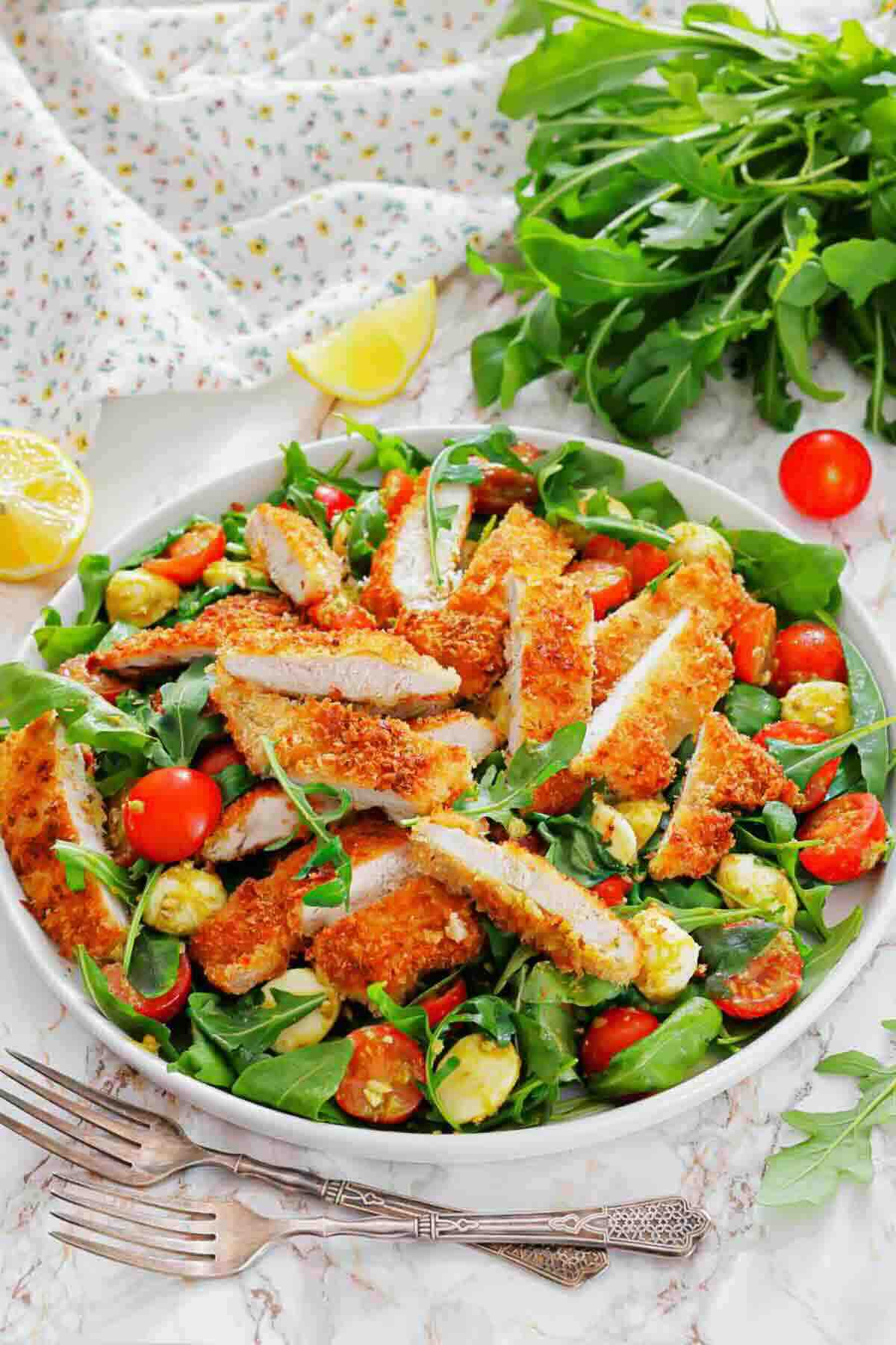 A bowl of salad with sliced breaded chicken, cherry tomatoes, mozzarella balls, and arugula, garnished with lemon wedges, on a marble surface with utensils and greens nearby.