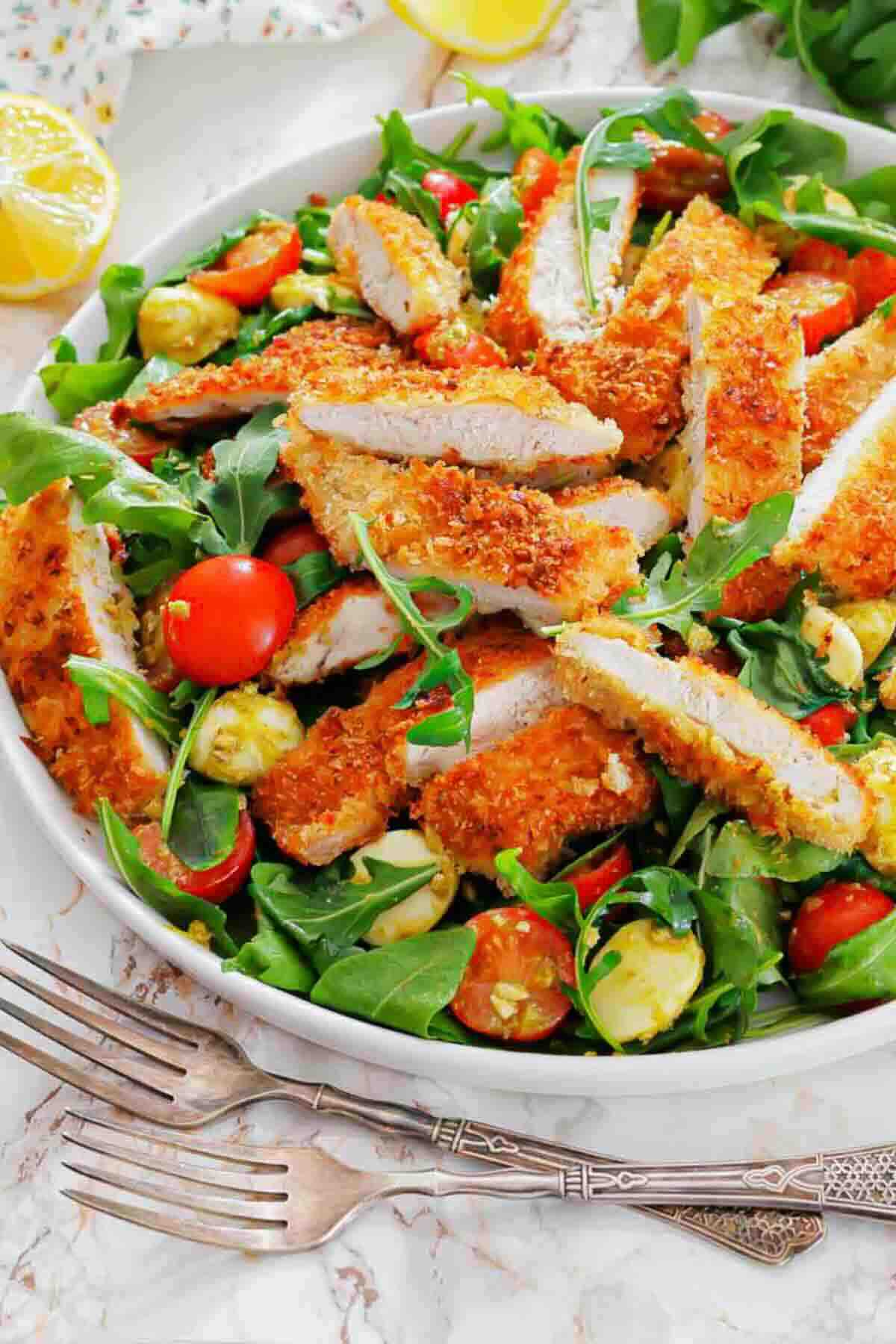 A salad with breaded chicken strips, cherry tomatoes, avocado pieces, and arugula served in a white bowl on a marble surface.