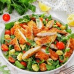 A salad with breaded chicken strips, cherry tomatoes, mozzarella balls, and arugula, garnished with lemon wedges.