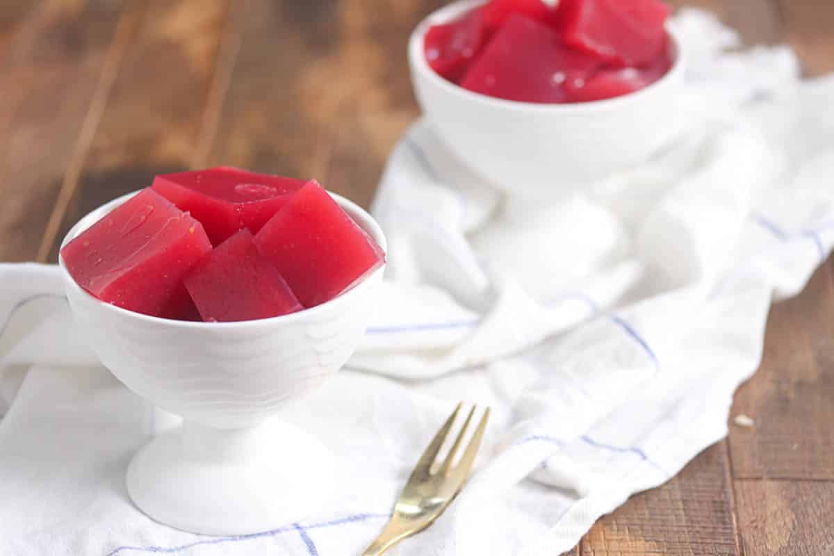 Two white dessert bowls filled with red gelatin cubes sit on a white cloth, with a gold fork placed in front.