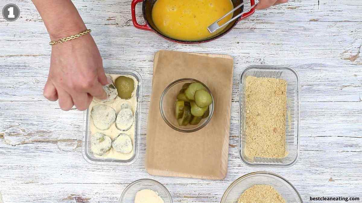 Person breading pickle slices by dipping them in flour, egg mixture, and breadcrumbs on a white wooden surface with ingredients arranged in glass dishes.