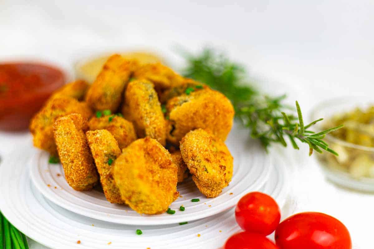 A white plate with golden-brown breaded pickles, garnished with herbs. Cherry tomatoes and bowls of sauce are in the background.