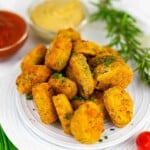 A plate of breaded and fried mushroom pieces, garnished with chopped chives, with two dipping sauces and fresh herbs in the background.