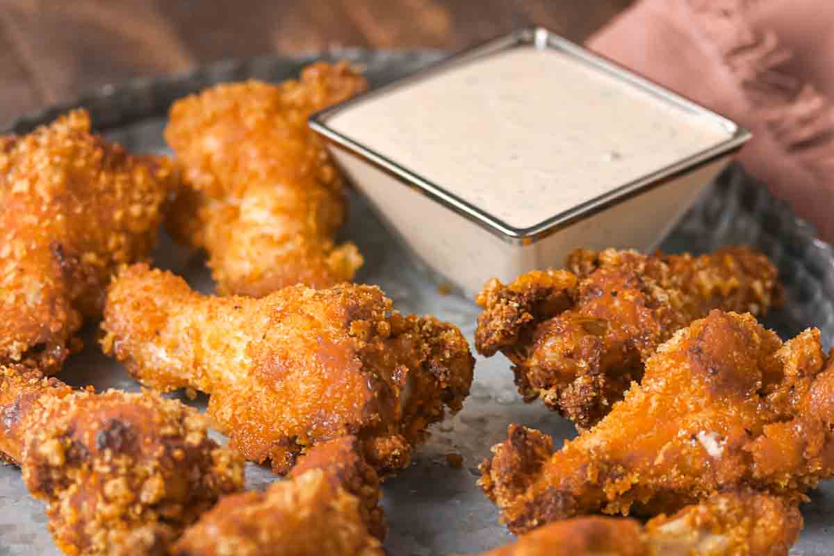 Fried chicken drumsticks arranged on a metal tray, served with a square dish of creamy dipping sauce.