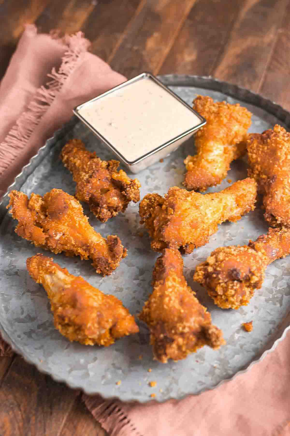 A round metal tray with seven crispy fried chicken wings arranged around a square dish of dipping sauce, placed on a wooden table with a pink napkin.