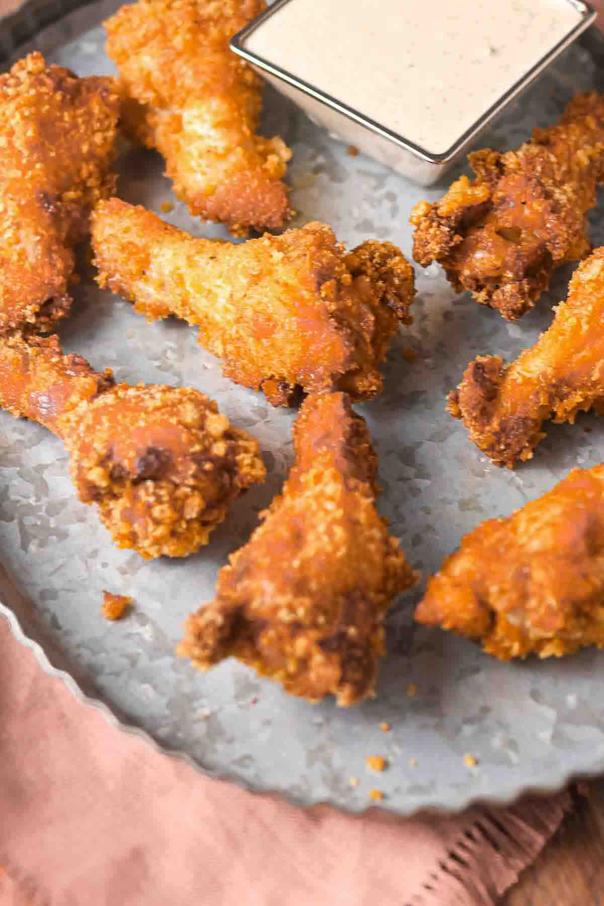 Several pieces of fried chicken wings are arranged on a metal tray with a small container of dipping sauce in the corner.