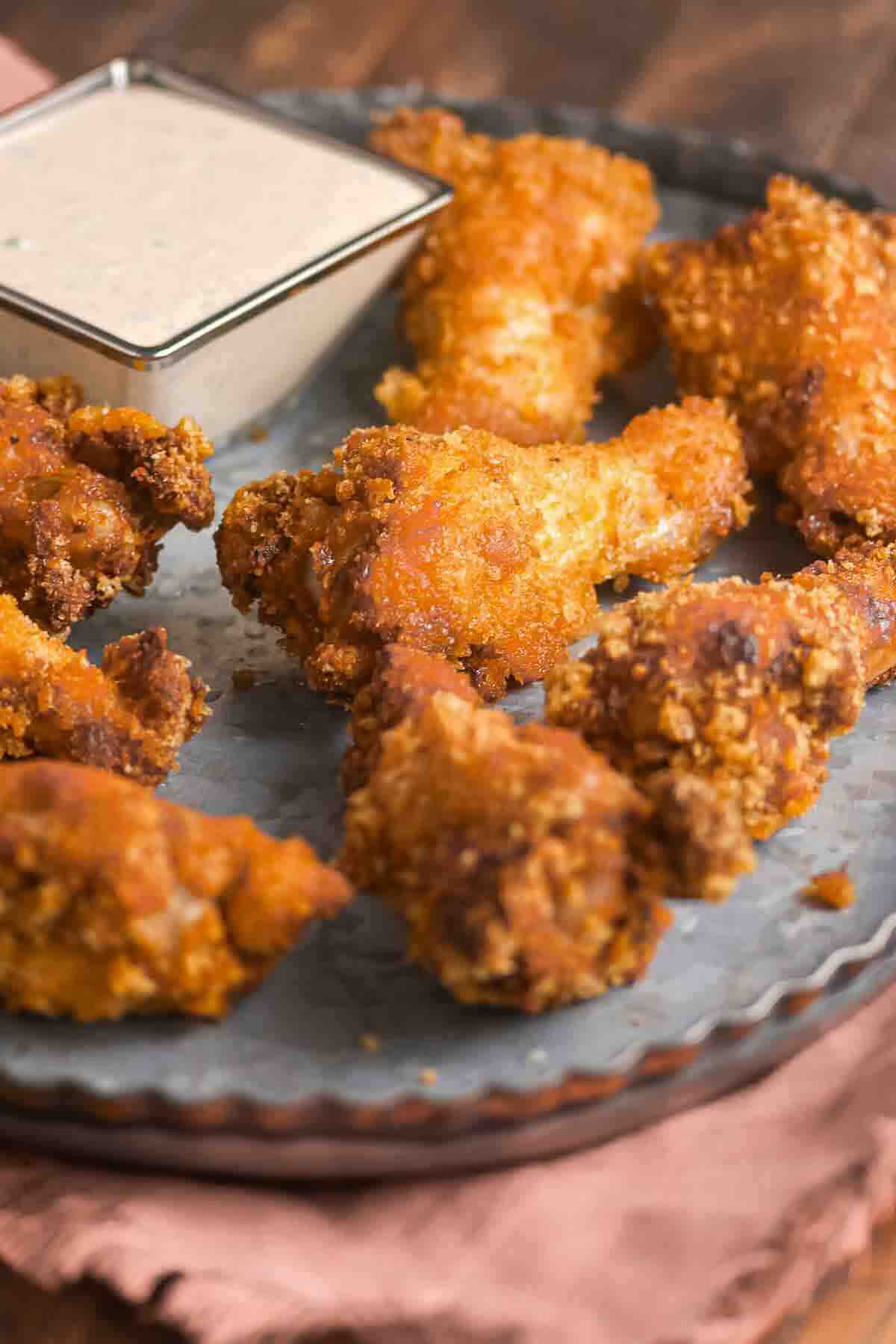 Eight pieces of crispy fried chicken wings on a round metal tray with a square dish of dipping sauce in the corner.