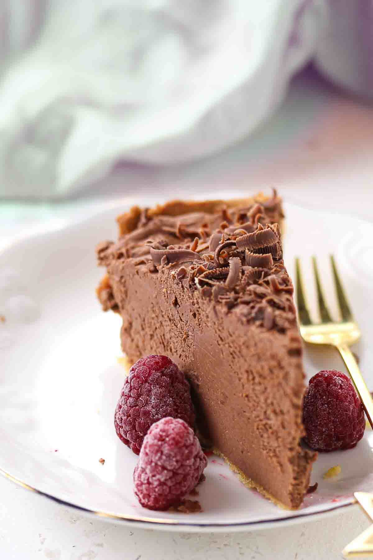 A slice of chocolate cheesecake garnished with chocolate shavings and three raspberries on a white plate with a gold fork.