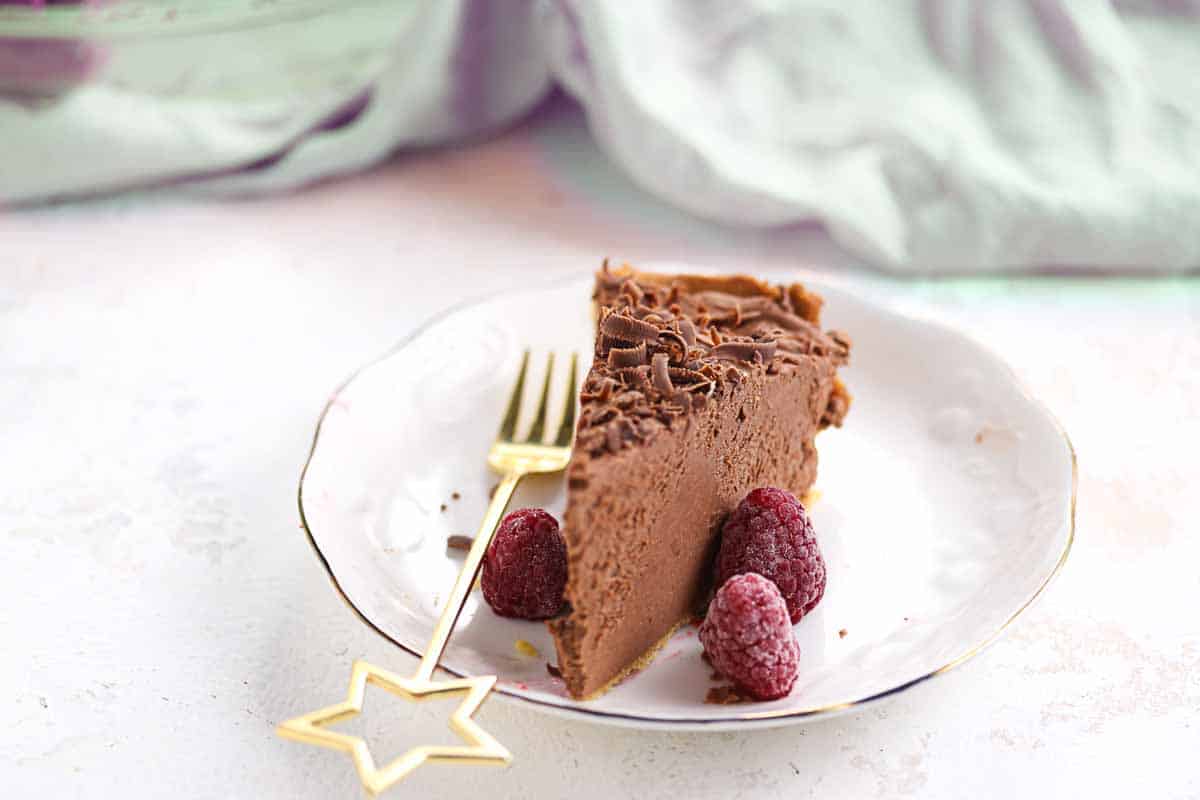 A slice of chocolate mousse pie with chocolate shavings and raspberries on a white plate, next to a gold fork with a star-shaped handle.