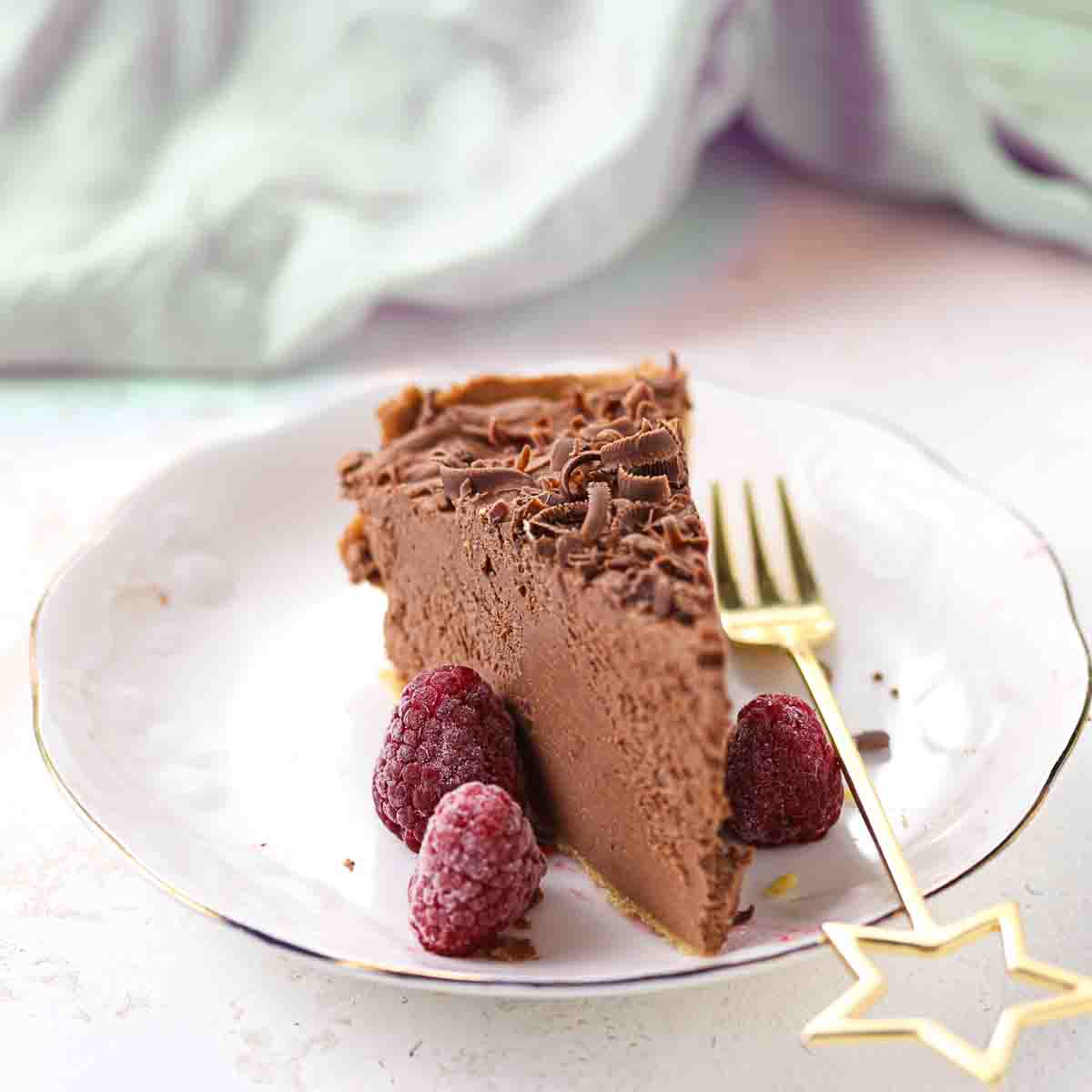 A slice of chocolate cheesecake on a white plate with three raspberries and a gold fork with a star-shaped handle.