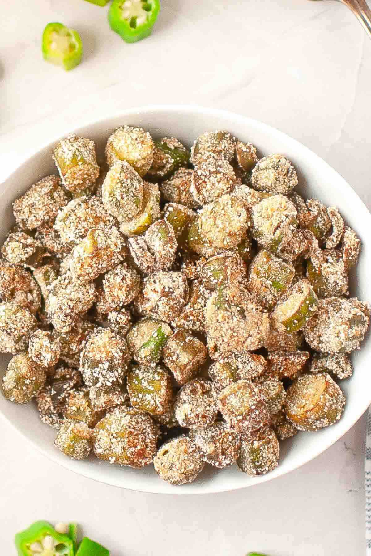 A white bowl filled with breaded and fried okra pieces, with some sliced green peppers visible nearby on a light background.