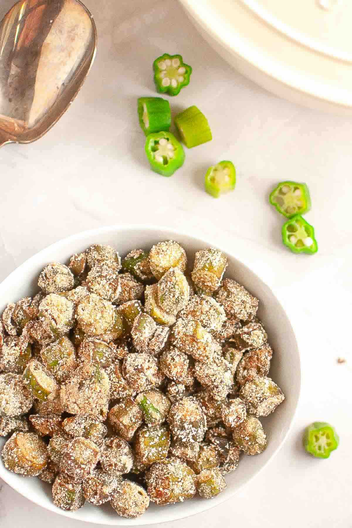 A white bowl filled with sliced okra coated in cornmeal sits on a marble surface, with a few raw okra slices scattered nearby.