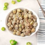 A white bowl filled with sliced, breaded, and fried okra sits on a light surface, with okra pieces scattered around and a striped towel nearby.