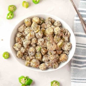 A white bowl filled with sliced, breaded, and fried okra sits on a light surface, with okra pieces scattered around and a striped towel nearby.