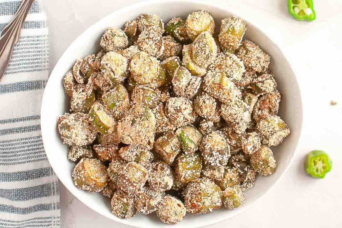 A white bowl filled with sliced, breaded, and fried okra pieces, sprinkled with seasoning, sits on a light surface near a striped towel.