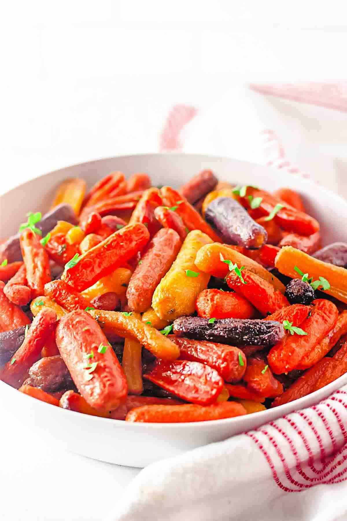 A white bowl filled with roasted baby carrots in various colors, garnished with small green herbs, sits on a white surface with a red-striped cloth nearby.