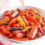 Bowl of roasted baby carrots in various colors, garnished with chopped herbs, placed on a white surface with a red-striped cloth nearby.