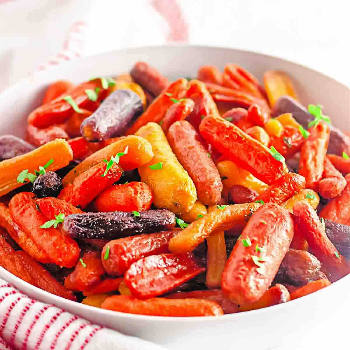 Bowl of roasted baby carrots in various colors, garnished with chopped herbs, placed on a white surface with a red-striped cloth nearby.