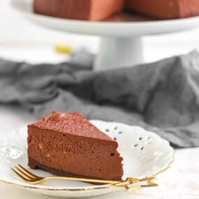 A slice of chocolate mousse cake on a white decorative plate with a gold fork; whole cake on a stand in the background.