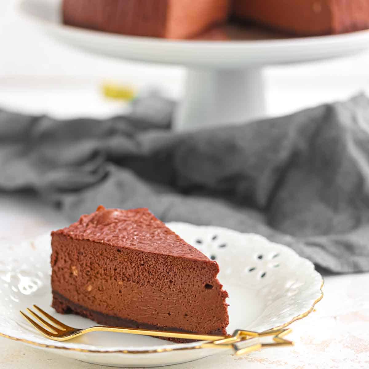 A slice of chocolate mousse cake on a white decorative plate with a gold fork; whole cake on a stand in the background.