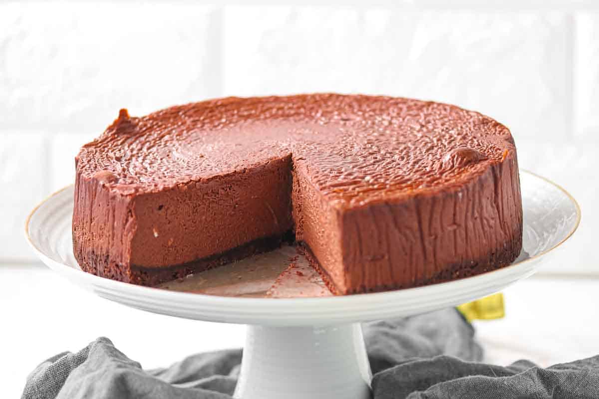 Chocolate cheesecake with one slice removed, displayed on a white cake stand against a light background.