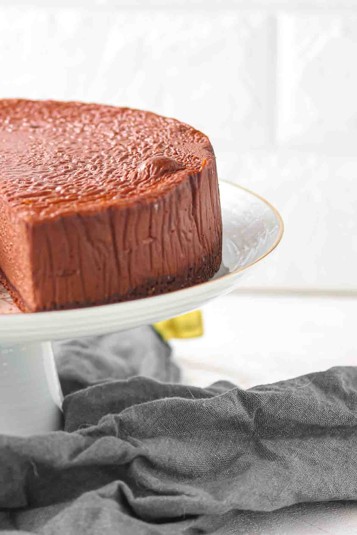 A chocolate cheesecake with a smooth top and textured sides sits on a white cake stand, partially sliced, with a gray cloth underneath.