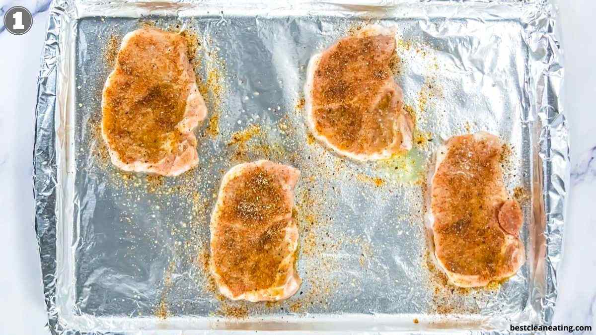 Four raw, seasoned pork chops are arranged on a foil-lined baking sheet, ready to be cooked.