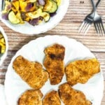 Five seasoned, cooked pork chops arranged on a white plate, with a separate plate of mixed roasted vegetables and two forks nearby on a wooden table.