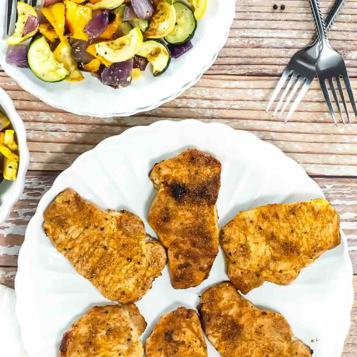 Five seasoned, cooked pork chops arranged on a white plate, with a separate plate of mixed roasted vegetables and two forks nearby on a wooden table.