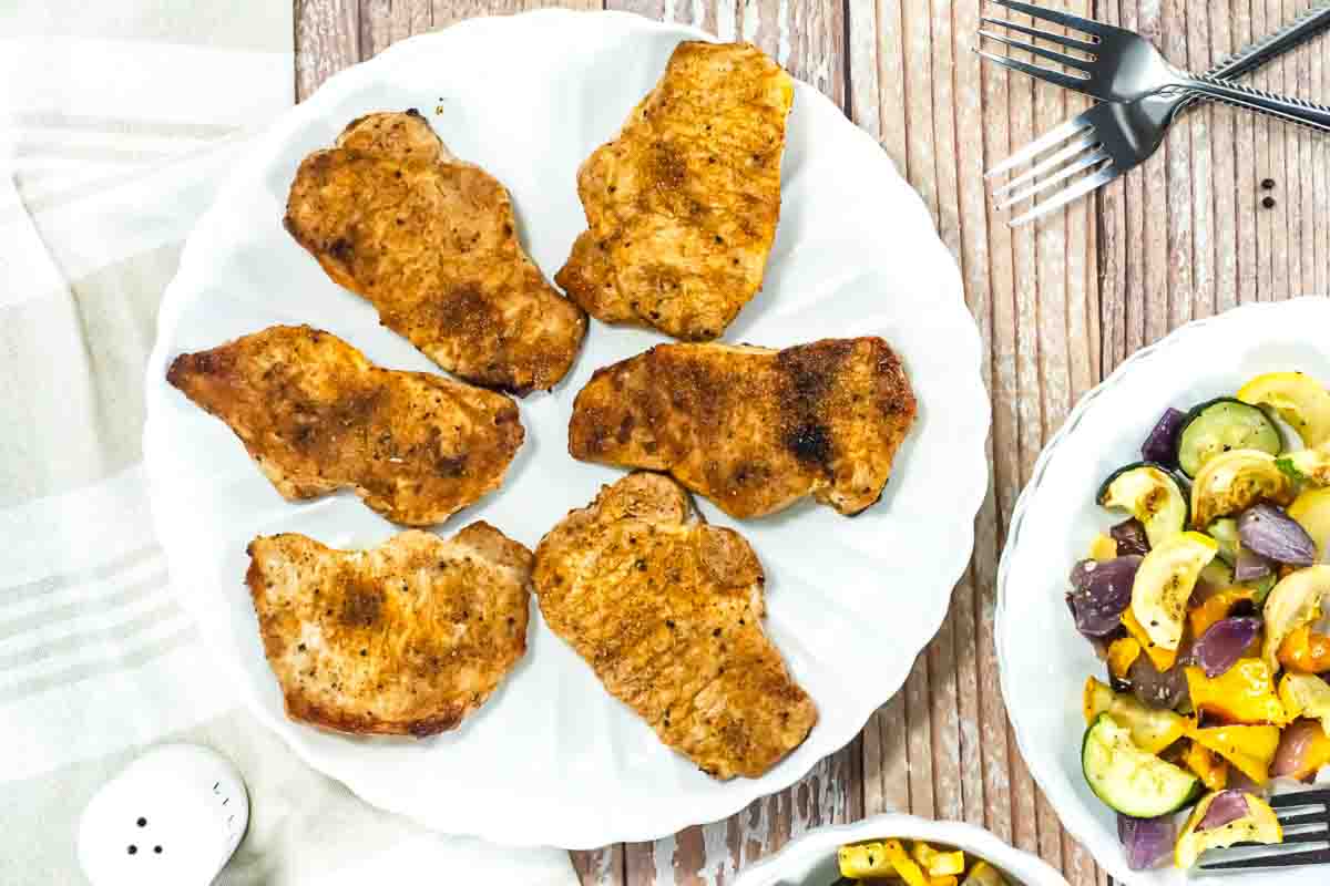 Six grilled pork chops arranged on a white plate, with a side dish of mixed roasted vegetables and two forks on a wooden table.