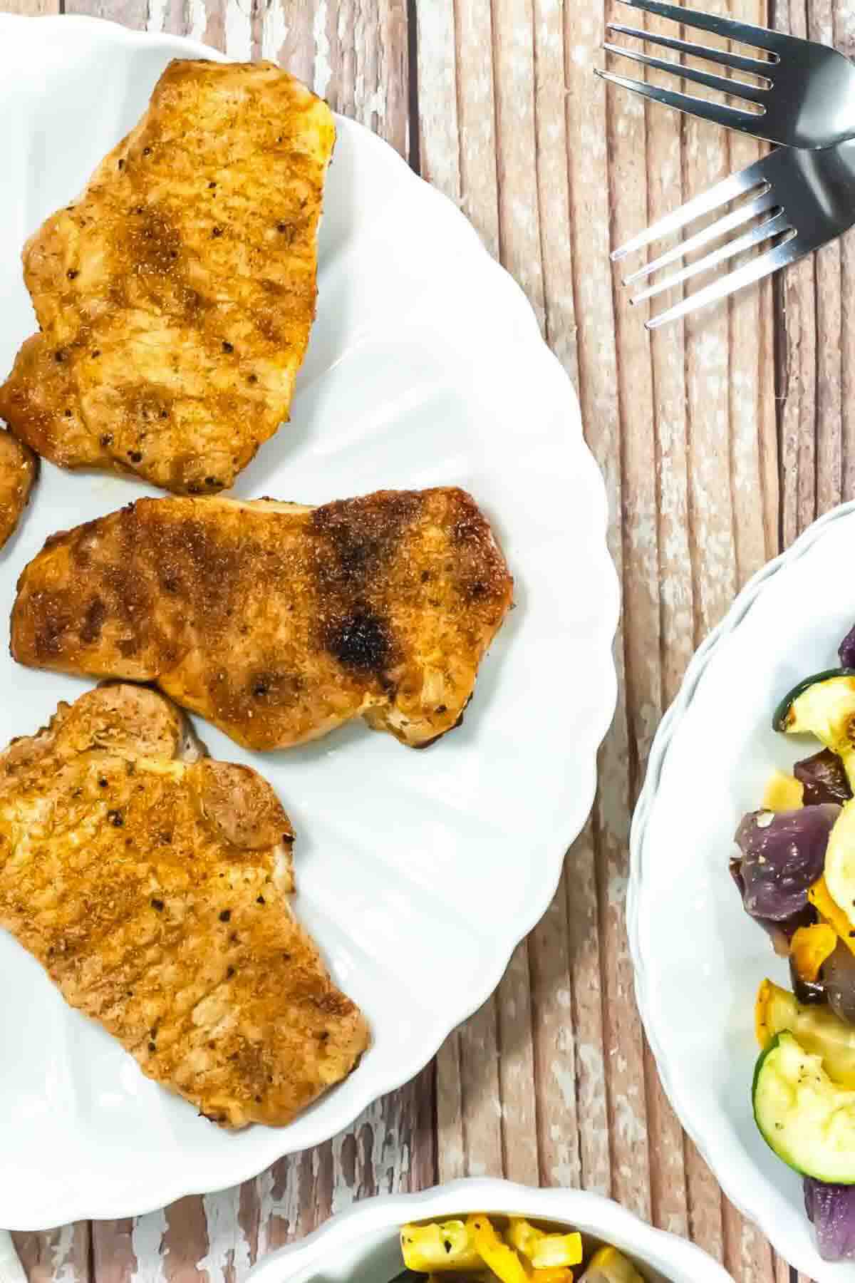 Three cooked pork chops on a white plate next to a dish of mixed roasted vegetables, placed on a wooden table with two forks nearby.