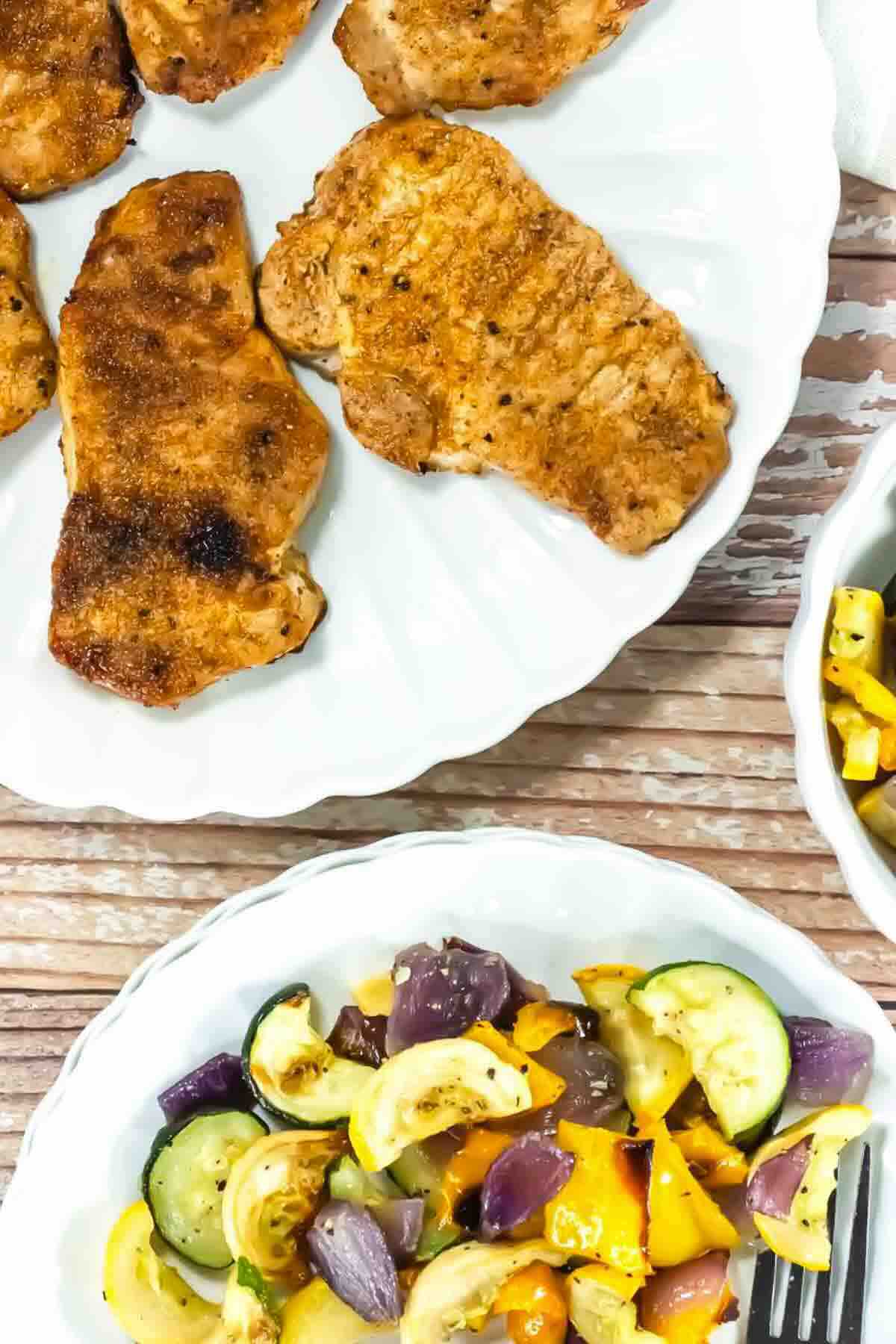 Grilled pork chops on a white plate, with a side of roasted mixed vegetables including zucchini, yellow squash, and red onion on another plate.