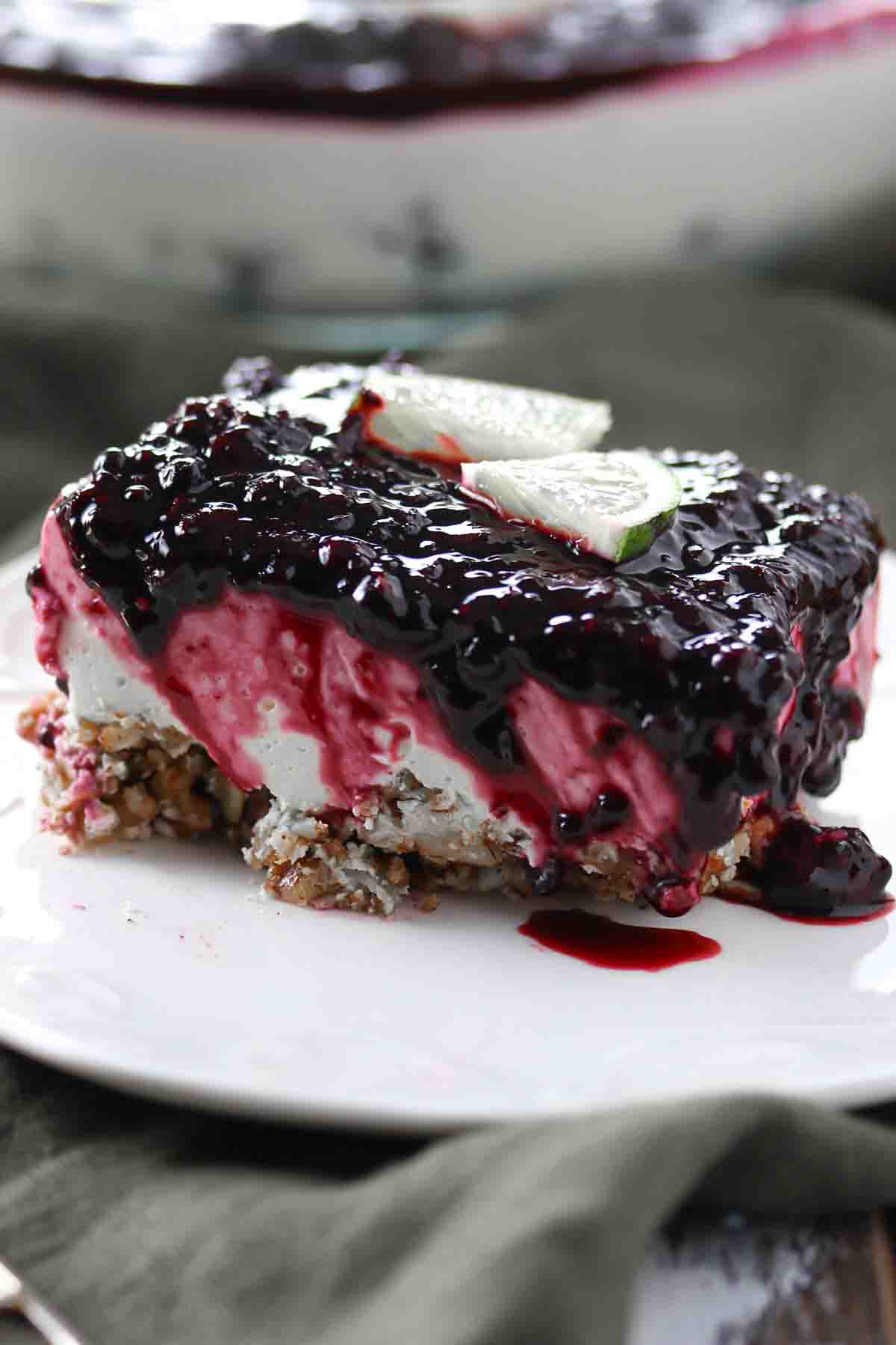 A slice of layered dessert with a nutty crust, creamy middle layer, and a blueberry topping, garnished with two lime slices on a white plate.