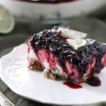 A slice of dessert with a nutty crust, creamy filling, and a layer of dark berry topping, garnished with two lime wedges, on a white plate.