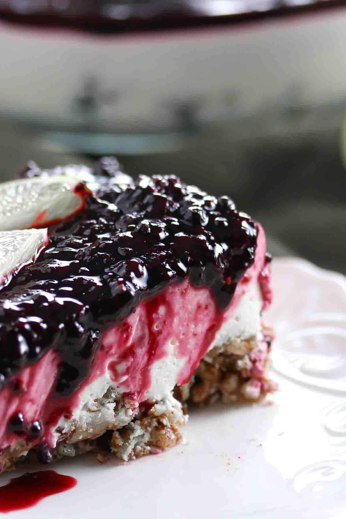 A close-up of a slice of dessert with a nutty crust, white and pink creamy layers, and a dark berry topping on a white plate.