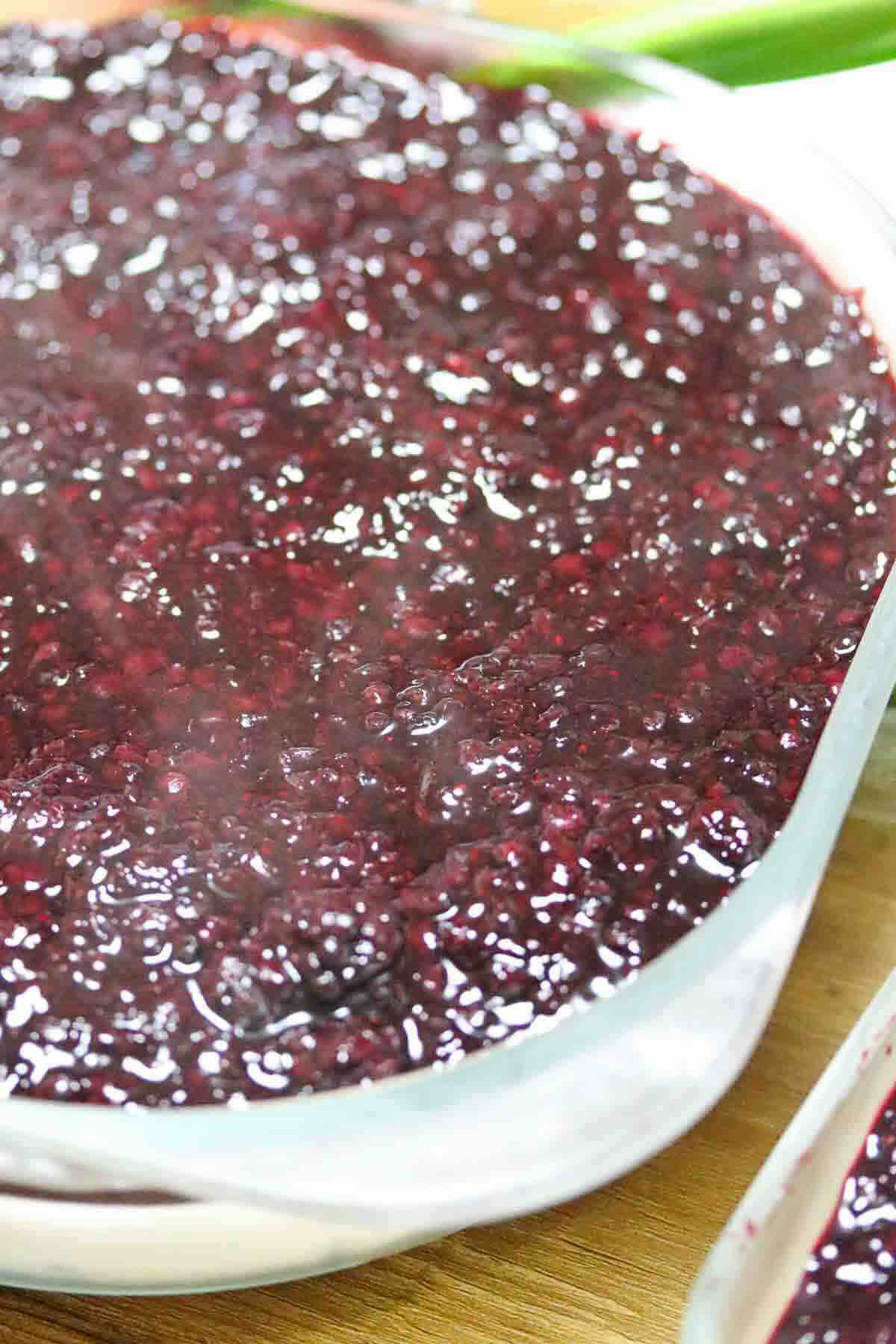 A close-up of a glass dish filled with thick, glossy, dark red berry compote on a wooden surface.