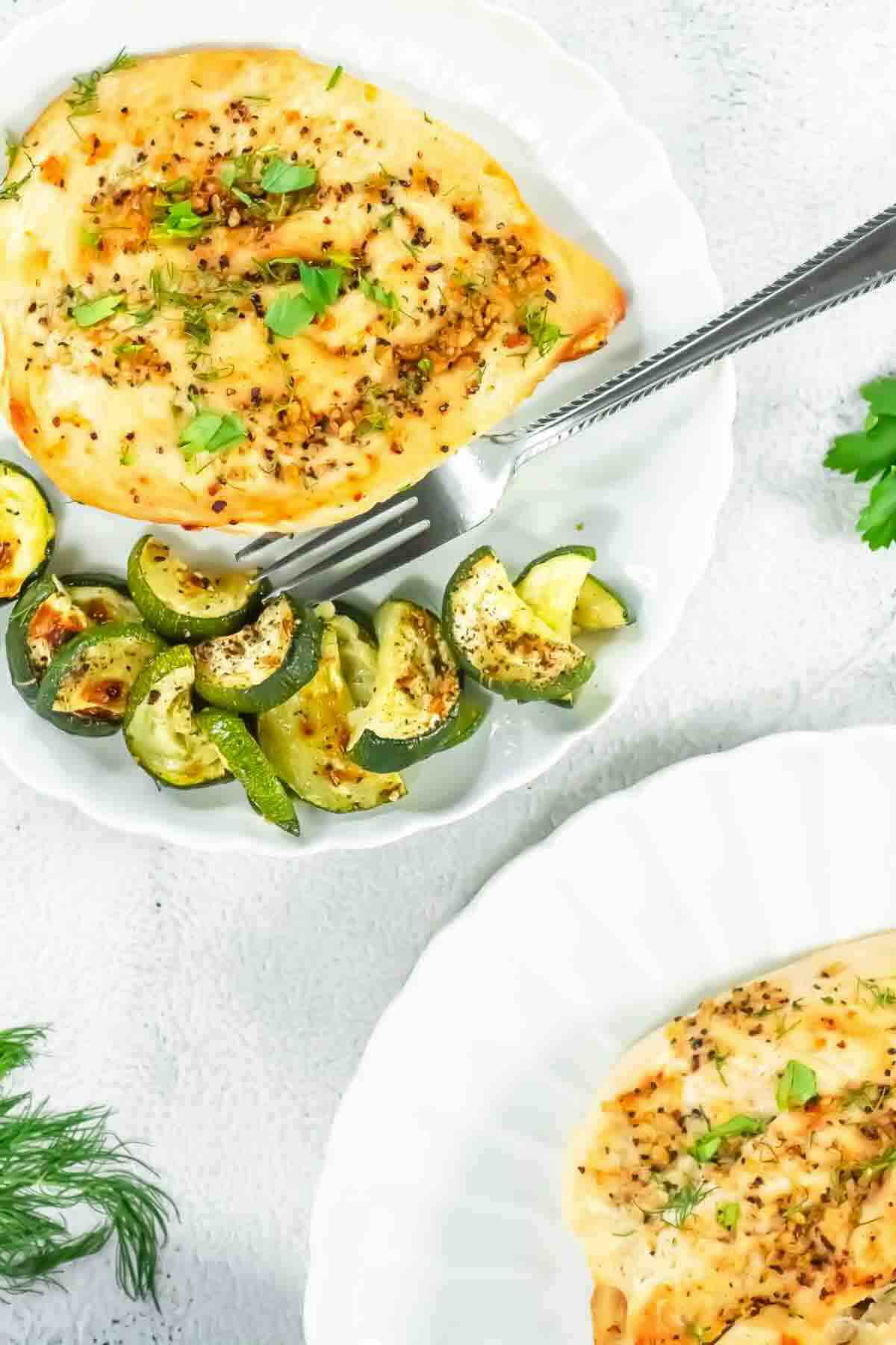 A plate with baked chicken breast topped with herbs, served with roasted zucchini slices, on a white surface with a fork.