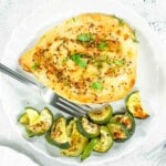 A cooked chicken breast garnished with herbs is served on a white plate alongside roasted zucchini pieces, with a fork resting on the plate.