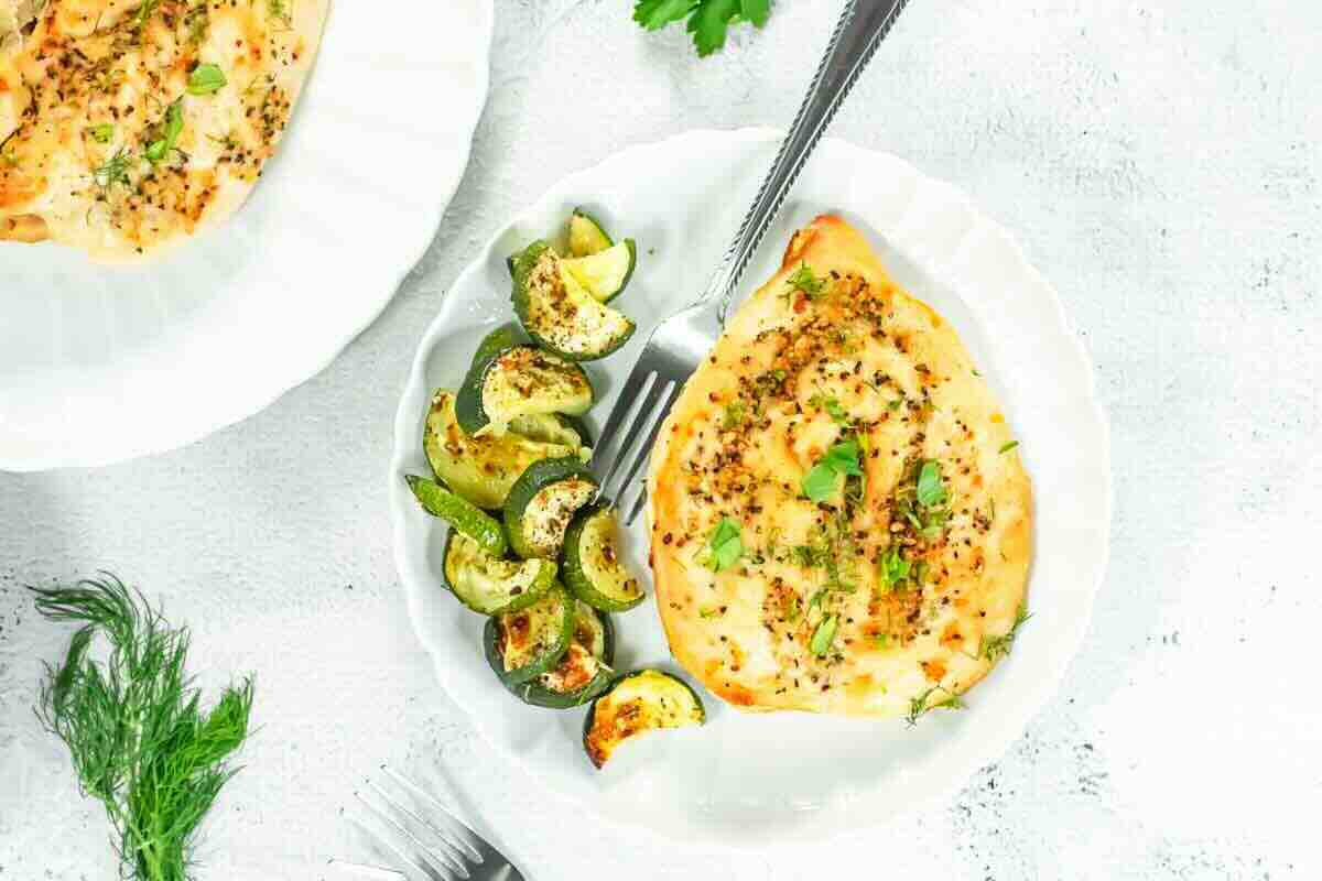 A white plate with baked chicken breast topped with herbs and melted cheese, served with roasted zucchini slices.