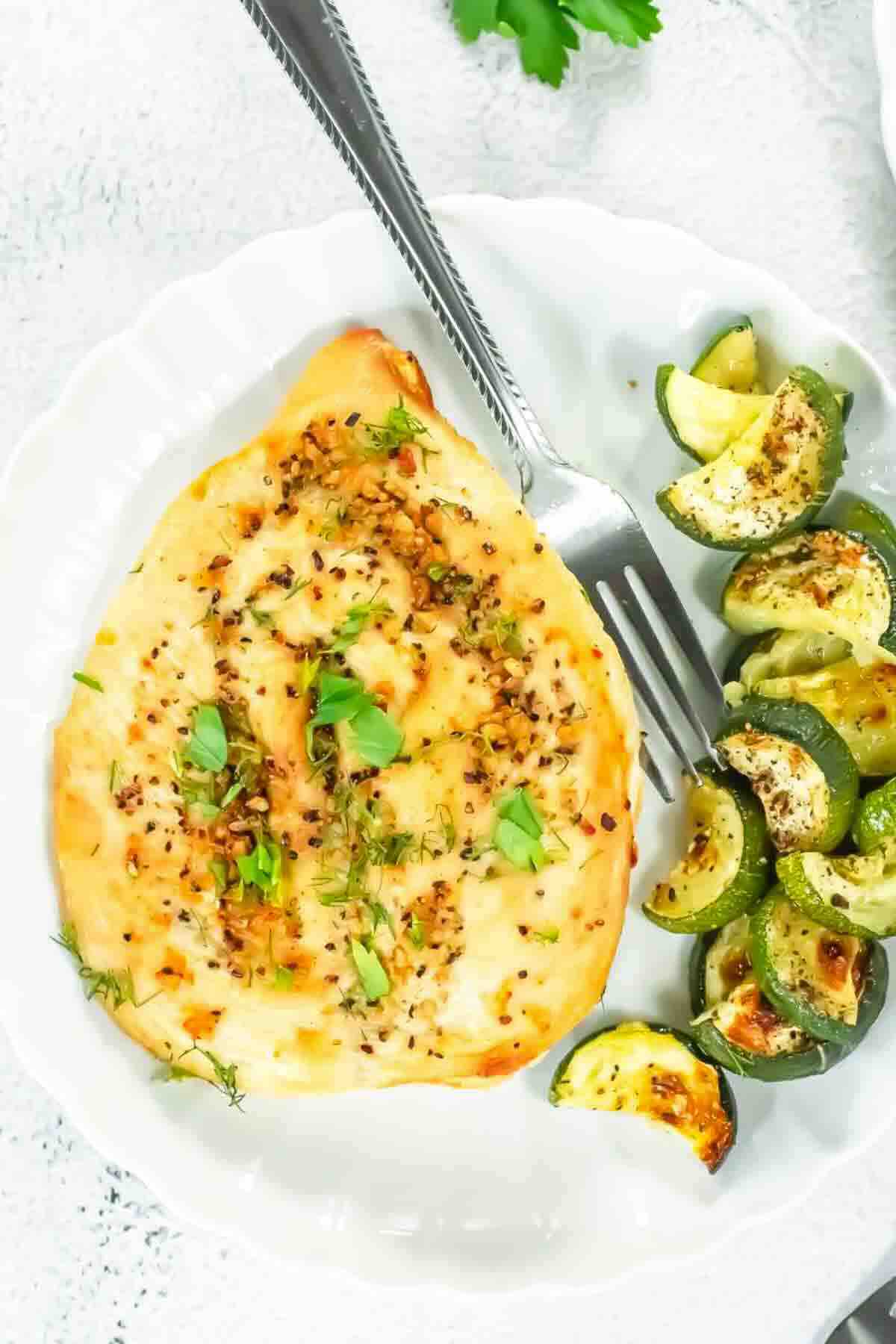Grilled chicken breast with herbs on a white plate, served with roasted zucchini slices and a fork.