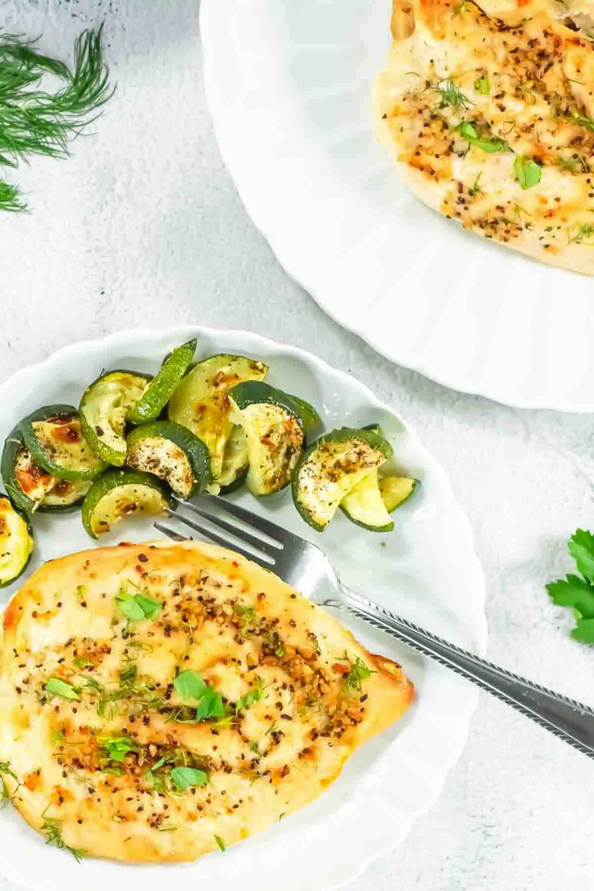 A white plate with baked chicken breast garnished with herbs and a side of roasted zucchini slices, with a fork placed on the plate.