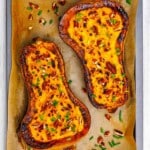 Two baked butternut squash halves topped with chopped pecans and chives on a parchment-lined baking sheet.