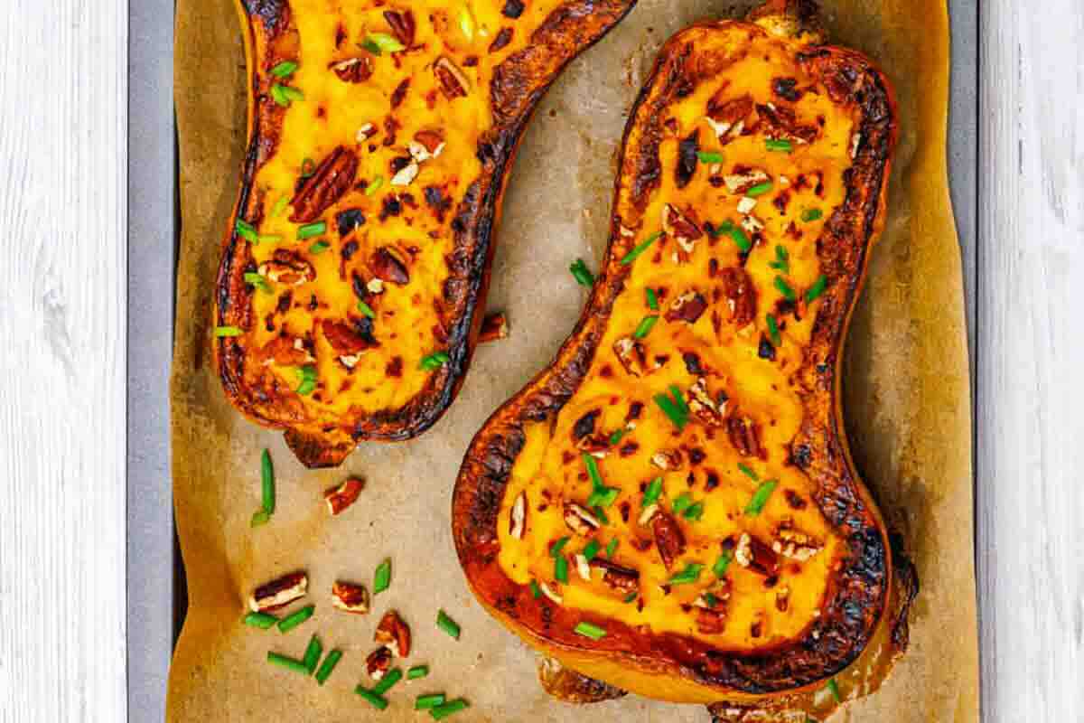 Two baked butternut squash halves filled with a creamy mixture, topped with chopped pecans and herbs, on a parchment-lined baking sheet.