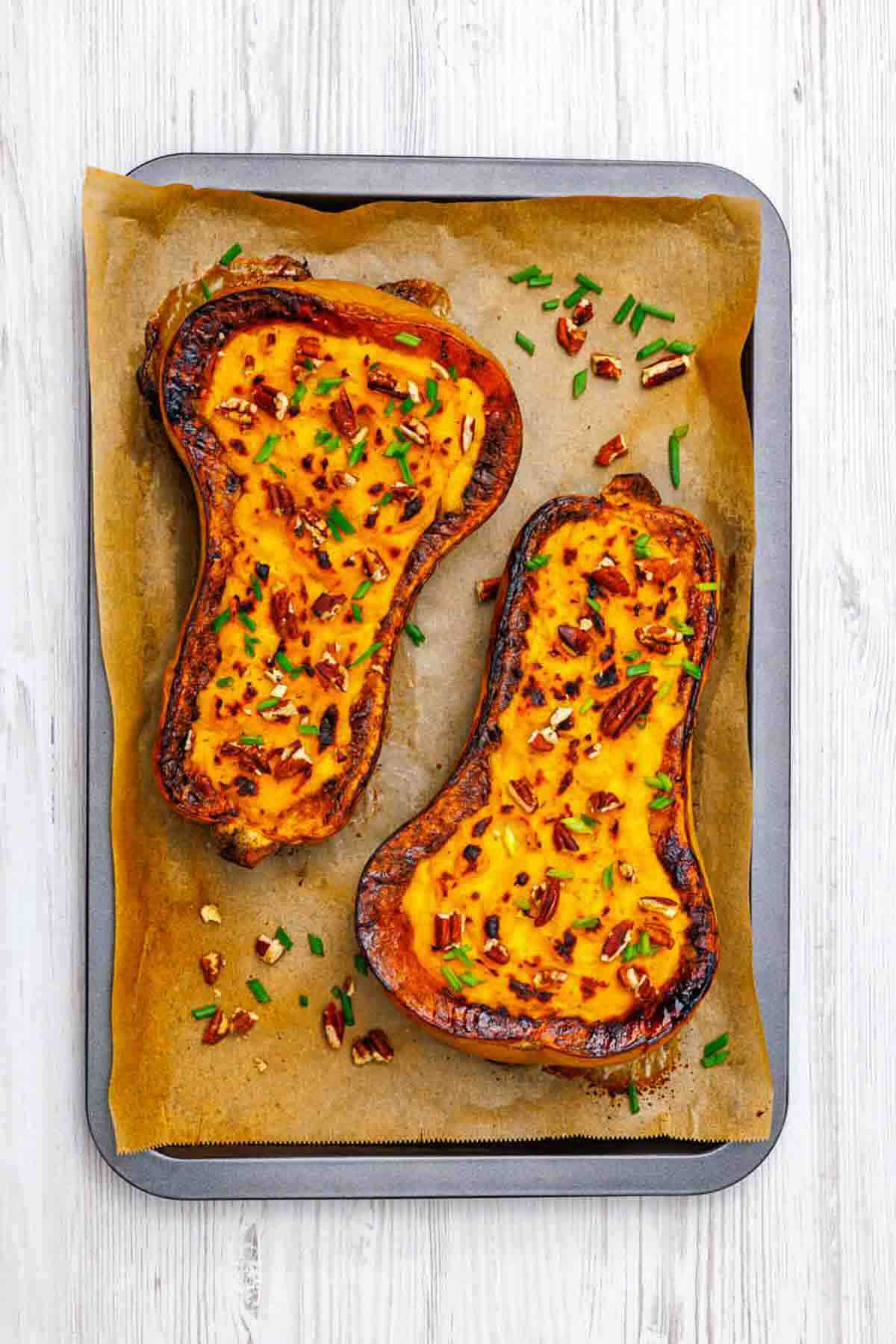 Two baked butternut squash halves filled with a cheesy mixture, garnished with chopped pecans and herbs, on a parchment-lined baking tray.