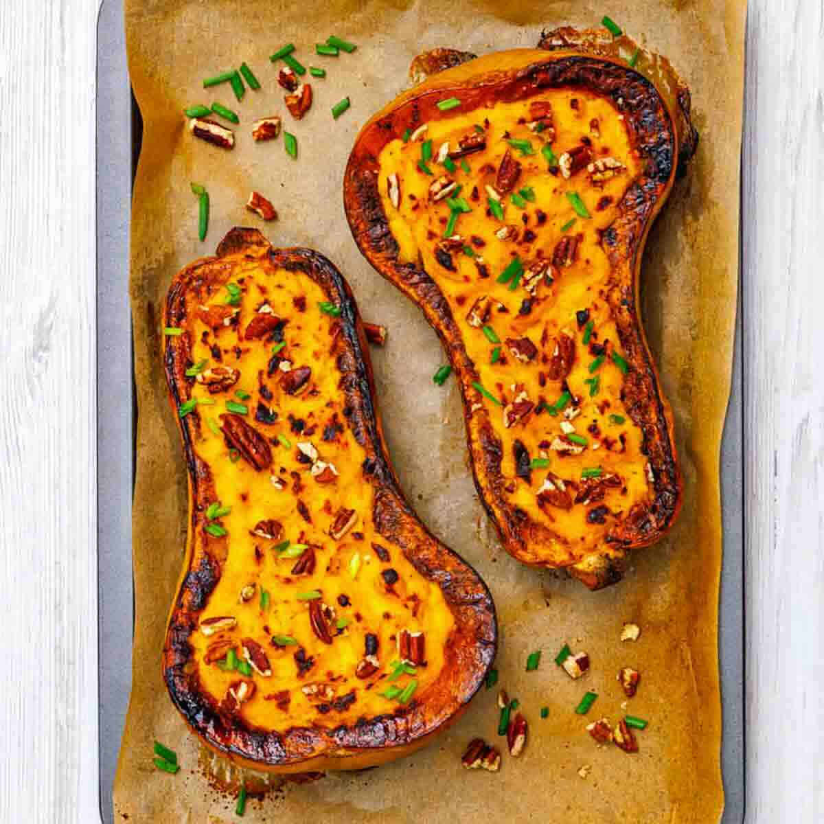 Two roasted butternut squash halves on parchment paper, topped with chopped pecans and green herbs.